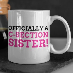 Peut inclure: Une tasse en céramique blanche avec un motif noir et rose. La tasse porte l'inscription "Officially a C-Section Sister!"