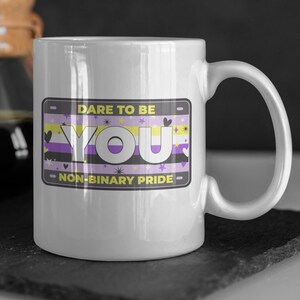 Könnte beinhalten: Weiße Keramiktasse mit einem Design aus einer gelben, violetten und weißen Non-Binary-Pride-Flagge. Die Tasse hat einen schwarzen Umriss und den Text "Dare to be You - Non-Binary Pride".