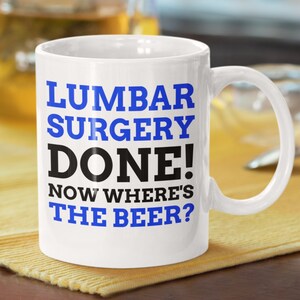 Puede incluir: Taza de café de cerámica blanca con texto azul y negro que dice "Cirugía lumbar realizada! Ahora, ¿dónde está la cerveza?"