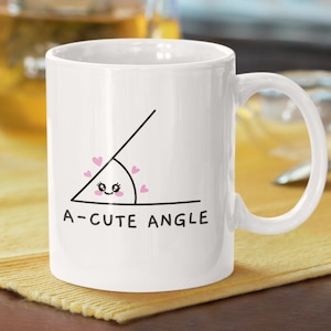May include: White ceramic mug featuring a black line drawing of a cute angle with pink hearts and the text "A-cute Angle".