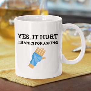 May include: White ceramic mug with a black graphic of a hand in a blue cast. The text on the mug reads "YES, IT HURT THANKS FOR ASKING."
