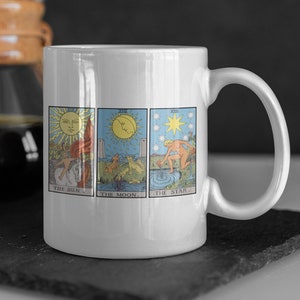 May include: White ceramic mug with a design of three tarot cards: The Sun, The Moon, and The Star. The cards are in a row, with the Sun card on the left, the Moon card in the middle, and the Star card on the right.