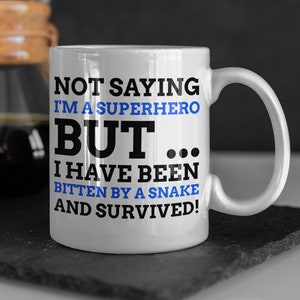 May include: White ceramic mug with a black and blue design. The mug reads "Not saying I'm a superhero but... I have been bitten by a snake and survived!"
