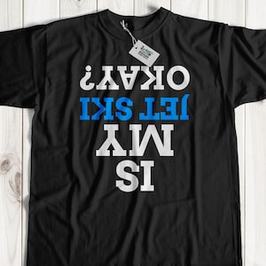 May include: Black t-shirt with the text "IS MY JET SKI OKAY?" in white and blue block letters. The shirt has a tag attached to the collar. The shirt is displayed on a light wood surface.