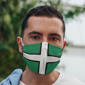 May include: A man wearing a white t-shirt and a face mask with the green and white flag of Cornwall. He is holding a smartphone in his right hand.