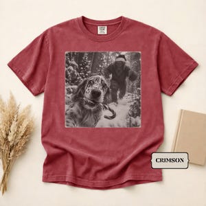 May include: A crimson short-sleeve t-shirt featuring a black and white graphic print of a dog holding a candy cane in its mouth with a Bigfoot figure walking in a snowy forest background. The shirt has a Comfort Colors brand label at the neck.
