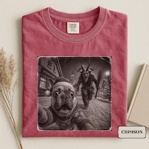 Könnte beinhalten: Ein karmesinrotes T-Shirt mit Rundhalsausschnitt und einem Grafikdruck einer Französischen Bulldogge mit Weihnachtsmütze und gemustertem Pullover, die ein Selfie mit einem gehörnten Krampus in einem verschneiten Dorf macht, Text Joyeux Noël.
