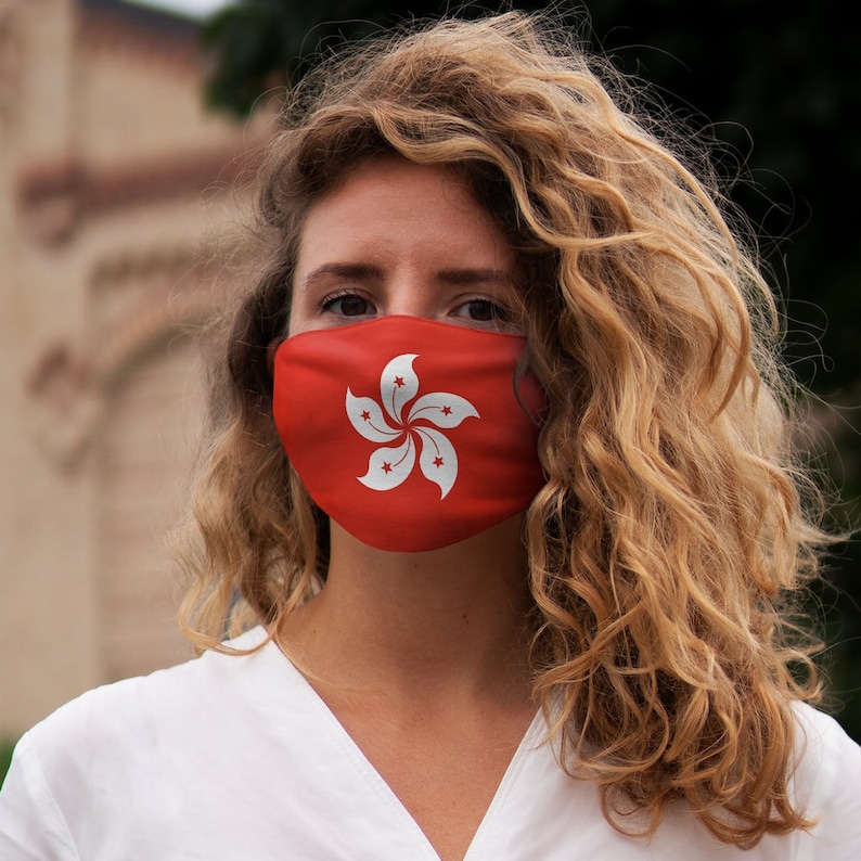 Op de afbeelding: Een vrouw met lang, krullend blond haar draagt ​​een rood mondkapje met de vlag van Hongkong erop.