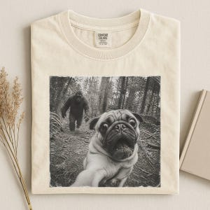 Pode incluir: Uma t-shirt de manga curta na cor creme com uma estampa gráfica em preto e branco de um pug tirando uma selfie numa floresta com um gorila caminhando ao fundo. A t-shirt tem uma etiqueta da marca Comfort Colors impressa na gola interna.