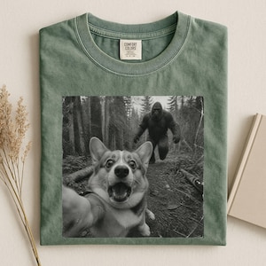 K&ouml;nnte beinhalten: Ein salbeigr&uuml;nes T-Shirt mit Rundhalsausschnitt und einem schwarz-wei&szlig;en Grafikdruck eines Corgis, der ein Selfie in einem Wald macht, w&auml;hrend im Hintergrund eine Bigfoot-Figur zu sehen ist. Das Comfort Colors Etikett ist im Kragen aufgedruckt.