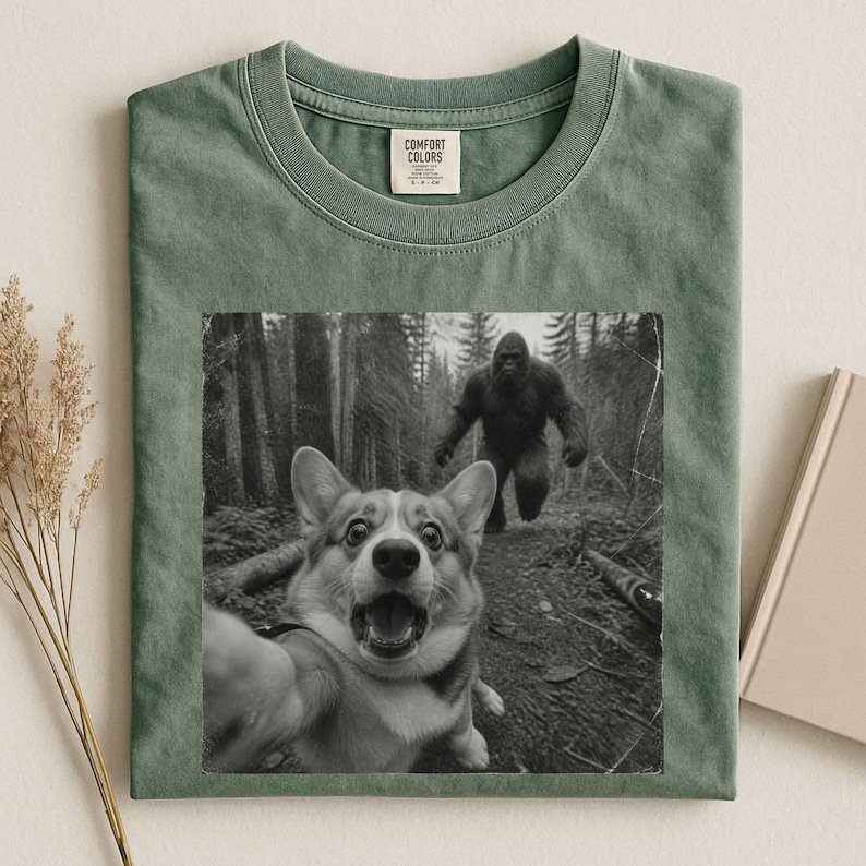 K&ouml;nnte beinhalten: Ein salbeigr&uuml;nes T-Shirt mit Rundhalsausschnitt und einem schwarz-wei&szlig;en Grafikdruck eines Corgis, der ein Selfie in einem Wald macht, w&auml;hrend im Hintergrund eine Bigfoot-Figur zu sehen ist. Das Comfort Colors Etikett ist im Kragen aufgedruckt.
