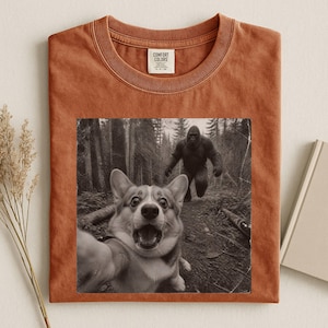 K&ouml;nnte beinhalten: Ein rostfarbenes T-Shirt mit Rundhalsausschnitt und einem schwarz-wei&szlig;en Grafikdruck, der einen Corgi beim Selfie in einem Wald zeigt, w&auml;hrend im Hintergrund eine Bigfoot-Figur zu sehen ist. Das Comfort Colors Etikett ist im Kragen aufgedruckt.