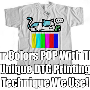 May include: White t-shirt featuring a cartoon cat with blue and white accents, holding a phone. Below the cat is a colorful test pattern. The shirt has the text "Our Colors POP With The UNIQUE DTG Printing Technique We Use!"