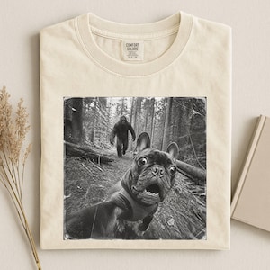 Könnte beinhalten: Ein cremefarbenes T-Shirt mit Rundhalsausschnitt und einem schwarz-weißen Grafikdruck, der eine Französische Bulldogge beim Selfie im Wald zeigt, mit einer Bigfoot-Figur im Hintergrund. Das Shirt hat ein Comfort Colors Etikett.