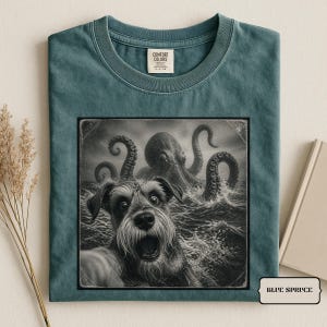 Op de afbeelding: Een blauw-sparrenkleurig t-shirt met een zwart-witte grafische print van een hond met een open bek op de voorgrond en een grote octopus die op de achtergrond uit de oceaangolven tevoorschijn komt. Het shirt heeft een Comfort Colors label.