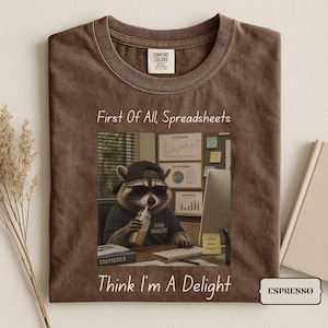 May include: A brown crew neck t-shirt featuring a graphic of a raccoon wearing a black baseball cap and sunglasses, drinking from a glass, with the text First Of All, Spreadsheets Think I'm A Delight and Data Analyst on the shirt.