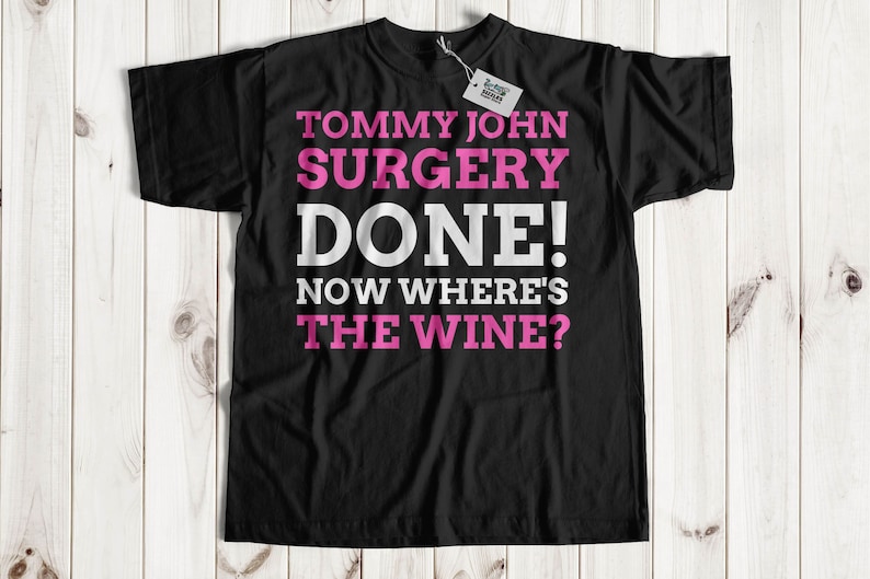 Puede incluir: Camiseta negra con el texto "TOMMY JOHN SURGERY DONE! NOW WHERE'S THE WINE?" en letras blancas y rosas. La camiseta tiene una etiqueta adjunta. La camiseta se muestra sobre un fondo de madera clara.