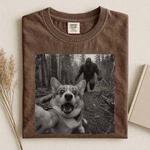 K&ouml;nnte beinhalten: Ein braunes T-Shirt mit Rundhalsausschnitt und einem Grafikdruck eines Corgis, der ein Selfie in einem Wald macht, w&auml;hrend im Hintergrund eine gro&szlig;e affenartige Kreatur zu sehen ist. Das Shirt hat ein Comfort Colors Etikett am Kragen.