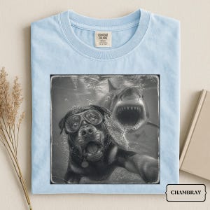 May include: A light blue t-shirt featuring a black and white graphic print of a Rottweiler wearing swimming goggles underwater, with a shark swimming behind it. The shirt has a crew neckline and a Comfort Colors brand label at the inner collar.