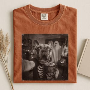 Könnte beinhalten: Ein rostfarbenes T-Shirt mit Rundhalsausschnitt und einem schwarz-weißen Grafikdruck einer Französischen Bulldogge, die ein Skelett-Shirt trägt und einen Eimer mit der Aufschrift Trick or Treat hält, mit einem Geist und einem Kürbis im Hintergrund.