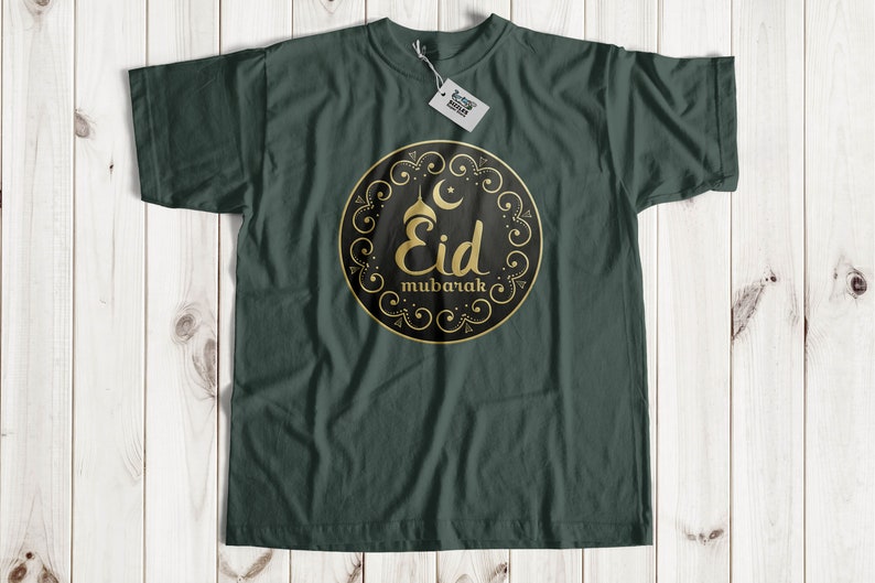 May include: Dark green t-shirt with a gold and black circular design. The design features the words "Eid Mubarak" in gold script, a crescent moon, and a star. The shirt is laid flat on a light wood surface.