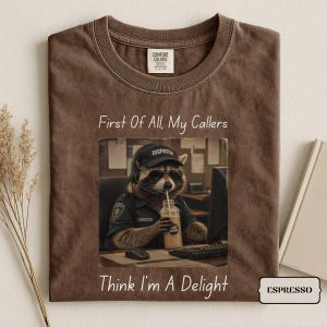 May include: A brown t-shirt featuring a graphic of a raccoon wearing a black 'DISPATCH' cap and sunglasses, sitting at a desk with a headset and iced coffee. The text 'First Of All, My Callers' appears above and 'Think I'm A Delight' below the graphic.