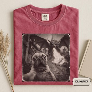 Peut inclure: Un t-shirt à manches courtes cramoisi avec une impression graphique en noir et blanc d'un bouledogue français prenant un selfie avec un dragon cracheur de feu en arrière-plan. Le t-shirt porte une étiquette Comfort Colors au col.