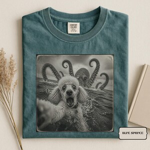 May include: A blue spruce coloured t-shirt with a crew neckline and a Comfort Colors label. The centre features a black and white graphic of a poodle with an open mouth in the ocean, with a large octopus tentacle creature emerging from the water.