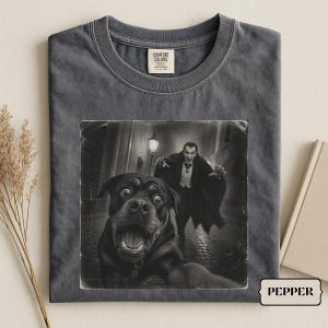 Könnte beinhalten: Ein anthrazitfarbenes T-Shirt mit Rundhalsausschnitt und einem Grafikdruck, der einen Rottweiler im Vordergrund und eine Vampirfigur im Hintergrund zeigt. Das Shirt hat ein Comfort Colors Markenetikett am Innenhalsausschnitt und ein kleines Etikett mit PEPPER.