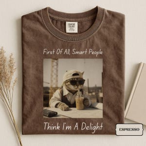 Puede incluir: Una camiseta marrón con un gráfico de un gato con gorra de béisbol y gafas de sol bebiendo de un vaso. El texto sobre el gato dice First Of All, Smart People y el texto debajo dice Think I'm A Delight. La camiseta tiene una etiqueta Comfort Colors.