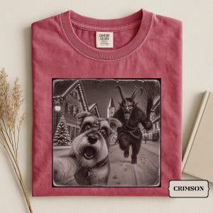 Peut inclure: Un t-shirt cramoisi avec une impression graphique représentant un schnauzer prenant un selfie avec une figure cornue de Krampus dans un village enneigé. Le t-shirt a un col rond et une étiquette de marque Comfort Colors à l'intérieur du col.
