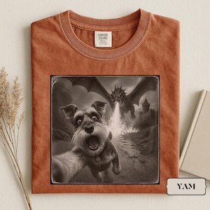 Op de afbeelding: Een roestkleurig T-shirt met ronde hals en een grafische print van een Schnauzer die een selfie maakt terwijl een draak op de achtergrond vuur spuwt. Het shirt heeft een Comfort Colors-label in de kraag en de tekst YAM op een klein label.