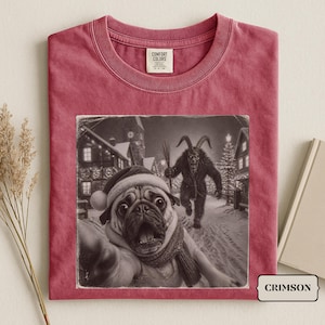 Może przedstawiać: Karmazynowy t-shirt z nadrukiem przedstawiającym mopsa w czapce Mikołaja i szaliku, robiącego selfie, podczas gdy postać Krampus zbliża się w zaśnieżonej wiosce. Koszulka ma okrągły dekolt i metkę marki Comfort Colors przy kołnierzu.