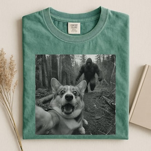 K&ouml;nnte beinhalten: Ein salbeigr&uuml;nes T-Shirt mit Rundhalsausschnitt und einem schwarz-wei&szlig;en Grafikdruck eines Corgis, der ein Selfie mit einer gro&szlig;en Sasquatch-Figur vor einem Waldhintergrund macht. Das Shirt hat ein Comfort Colors Markenetikett am Innenkragen.