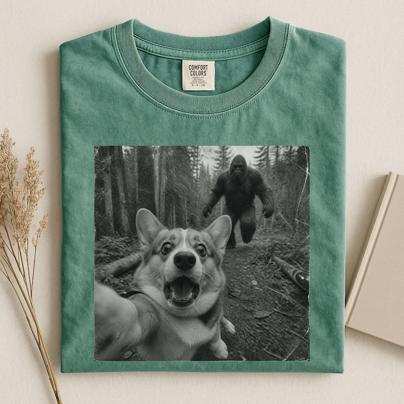 K&ouml;nnte beinhalten: Ein salbeigr&uuml;nes T-Shirt mit Rundhalsausschnitt und einem schwarz-wei&szlig;en Grafikdruck eines Corgis, der ein Selfie mit einer gro&szlig;en Sasquatch-Figur vor einem Waldhintergrund macht. Das Shirt hat ein Comfort Colors Markenetikett am Innenkragen.