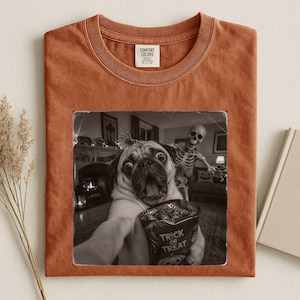 Pode incluir: Uma t-shirt de gola redonda em cor laranja queimado com uma estampa gráfica a preto e branco de um pug a tirar uma selfie com um esqueleto numa sala. O pug segura um saco de doces com a etiqueta Trick or Treat. Etiqueta da marca Comfort Colors.