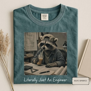 Puede incluir: Una camiseta color abeto azul con un gráfico de un mapache con gorra y gafas de sol, sosteniendo una bebida mientras trabaja en un escritorio con planos. El texto debajo del gráfico dice, Literally Just An Engineer.