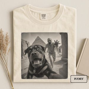 Könnte beinhalten: Ein elfenbeinfarbenes T-Shirt mit Rundhalsausschnitt und einem Grafikdruck, der einen Rottweiler im Vordergrund zeigt, während eine Mumie im Hintergrund in der Nähe ägyptischer Pyramiden hinterherläuft. Das Etikett Comfort Colors ist am Kragen.