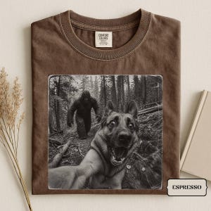以下が含まれることがあります： 茶色のクルーネックTシャツ。森の中で自撮りをするジャーマンシェパードと、背景を歩くビッグフットの白黒グラフィックプリントが施されています。首元にはComfort Colorsのブランドラベルが付いています。