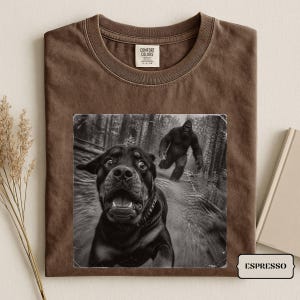 May include: A brown crew neck t-shirt featuring a black and white graphic print of a startled Rottweiler dog running away from a Bigfoot creature in a forest. The shirt has a Comfort Colors brand label on the inside collar and the word ESPRESSO.