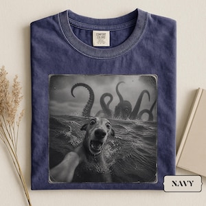 Può includere: Una t-shirt blu navy con una stampa grafica in bianco e nero di un cane che scatta un selfie nell'oceano mentre un grande polpo emerge dall'acqua dietro di lui. La maglietta ha un girocollo e un'etichetta del marchio Comfort Colors.