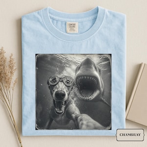 May include: A light blue t-shirt featuring a black and white graphic print of a dog wearing goggles underwater with a shark swimming behind it. The shirt has a crew neckline and a Comfort Colors brand label printed on the inside of the collar.