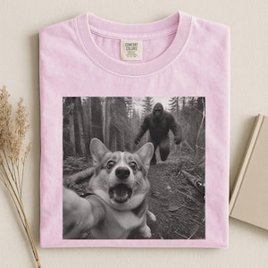 K&ouml;nnte beinhalten: Ein hellrosa T-Shirt mit einem schwarz-wei&szlig;en Grafikdruck eines Corgi-Hundes, der ein Selfie in einem Wald macht, w&auml;hrend im Hintergrund eine gro&szlig;e Bigfoot-Kreatur l&auml;uft. Das T-Shirt hat ein Comfort Colors Markenetikett im inneren Nackenbereich.