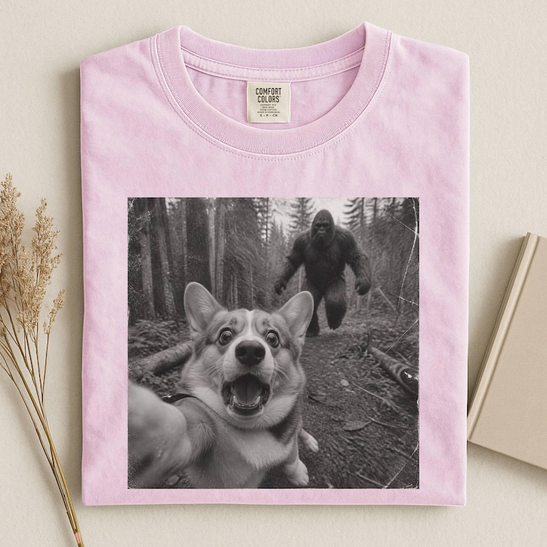 K&ouml;nnte beinhalten: Ein hellrosa T-Shirt mit einem schwarz-wei&szlig;en Grafikdruck eines Corgi-Hundes, der ein Selfie in einem Wald macht, w&auml;hrend im Hintergrund eine gro&szlig;e Bigfoot-Kreatur l&auml;uft. Das T-Shirt hat ein Comfort Colors Markenetikett im inneren Nackenbereich.
