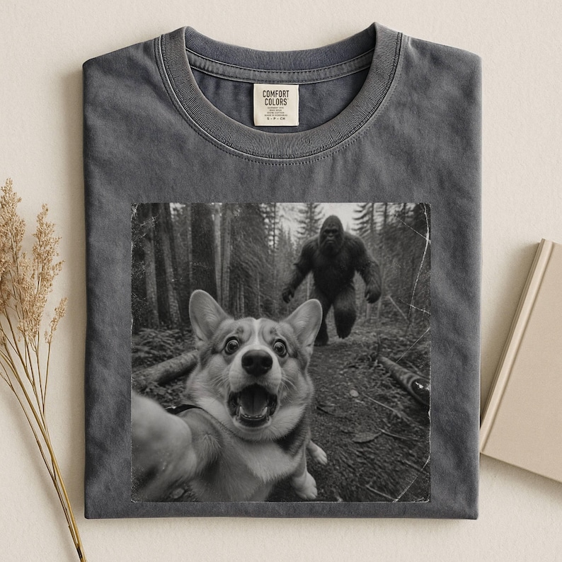 K&ouml;nnte beinhalten: Ein anthrazitfarbenes T-Shirt mit Rundhalsausschnitt und einem schwarz-wei&szlig;en Grafikdruck eines Corgis, der ein Selfie im Wald macht, w&auml;hrend im Hintergrund eine Bigfoot-Figur erscheint. Das Etikett Comfort Colors befindet sich am Innenkragen.