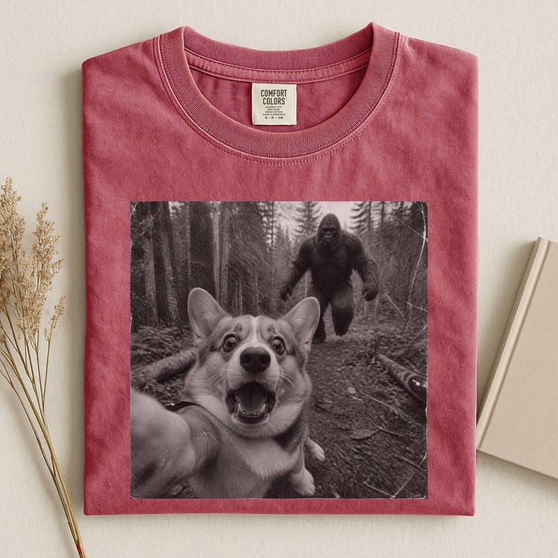 K&ouml;nnte beinhalten: Ein dunkelrotes T-Shirt mit Rundhalsausschnitt und einem schwarz-wei&szlig;en Grafikdruck eines Corgis, der ein Selfie in einem Wald macht, w&auml;hrend im Hintergrund eine Bigfoot-Figur zu sehen ist. Das Comfort Colors Etikett ist am Kragen.