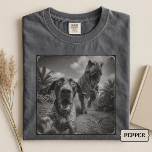 Op de afbeelding: Een antracietgrijs t-shirt met een zwart-witte grafische print van een hond die een selfie maakt met een Tyrannosaurus Rex in een jungle. Het shirt heeft een ronde hals en een Comfort Colors merklabel aan de binnenkant van de kraag.