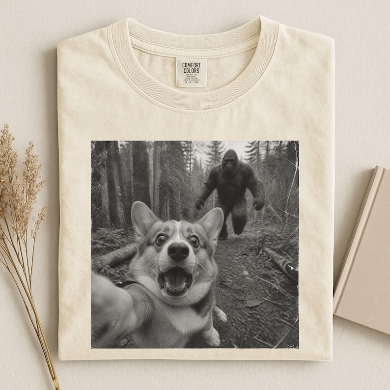 K&ouml;nnte beinhalten: Ein cremefarbenes T-Shirt mit einem schwarz-wei&szlig;en Grafikdruck eines Corgis, der ein Selfie im Wald macht, w&auml;hrend im Hintergrund eine Bigfoot-Figur l&auml;uft. Das T-Shirt hat einen Rundhalsausschnitt und ein Comfort Colors Etikett am Innenkragen.