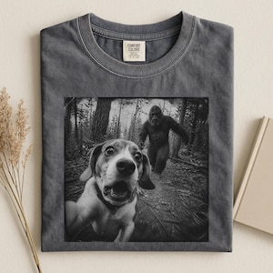 Könnte beinhalten: Ein anthrazitfarbenes T-Shirt mit Rundhalsausschnitt und einem schwarz-weißen Grafikdruck, der einen Beagle mit überraschtem Gesichtsausdruck im Vordergrund und eine große, haarige Kreatur, die Bigfoot ähnelt, im Wald zeigt.