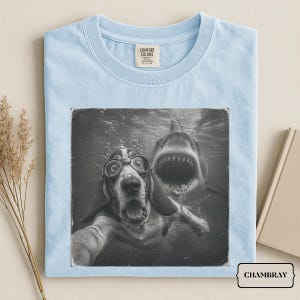May include: A light blue t-shirt featuring a black and white graphic print of a dog wearing goggles swimming underwater with a shark behind it. The shirt has a crew neckline and a Comfort Colors brand label printed on the inside of the collar.